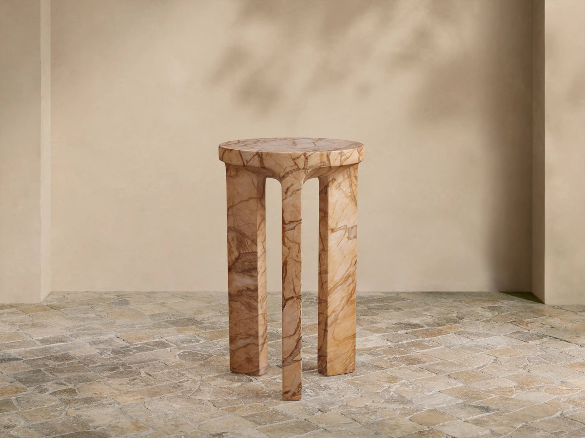 Odessa Outdoor End Table. Cylindrical top, three thick, curved legs, light tan with dark brown stone-like veining, on tiled floor.