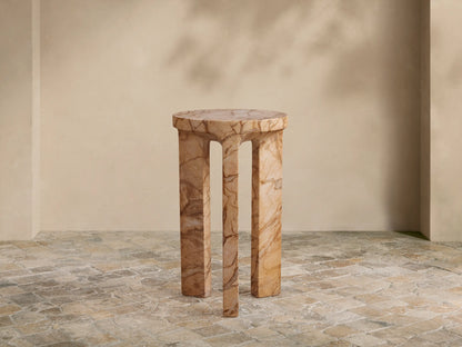 Odessa Outdoor End Table. Cylindrical top, three thick, curved legs, light tan with dark brown stone-like veining, on tiled floor.