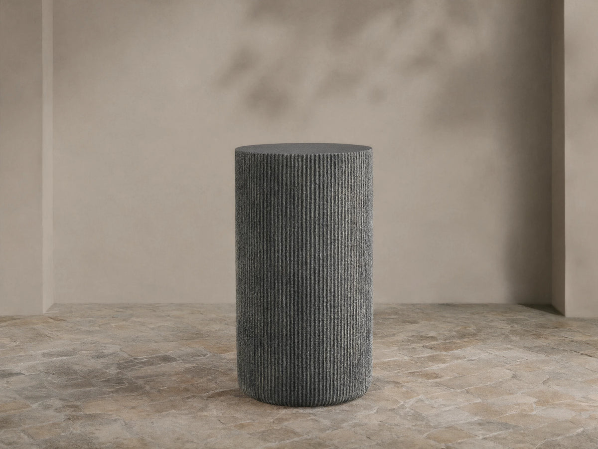 Ojai Outdoor End Table, dark gray, cylindrical with vertical ribbed texture, on a stone patio against a light beige wall.