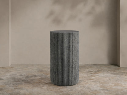 Ojai Outdoor End Table, dark gray, cylindrical with vertical ribbed texture, on a stone patio against a light beige wall.