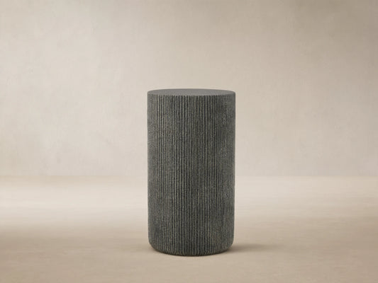 Ojai Outdoor End Table, dark gray, cylindrical with vertical ribbed texture, on a stone patio against a light beige wall.