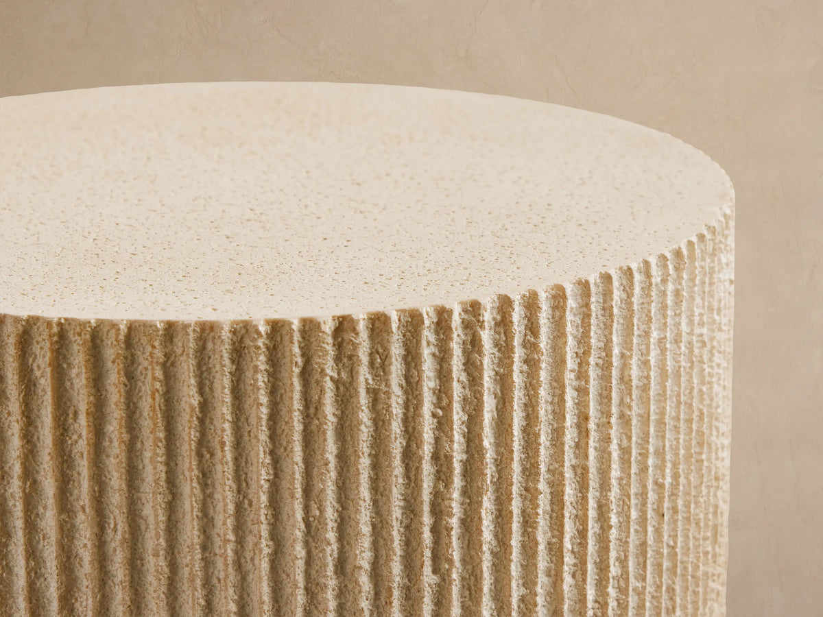 Front view of the light natural stone Ojai Outdoor End Table, cylindrical with a subtle ribbed texture, next to a wicker armchair.