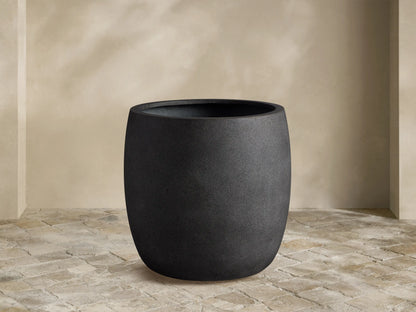 Textured, dark gray Ojai Outdoor Planter, barrel-shaped, on a light stone patio against a beige wall.