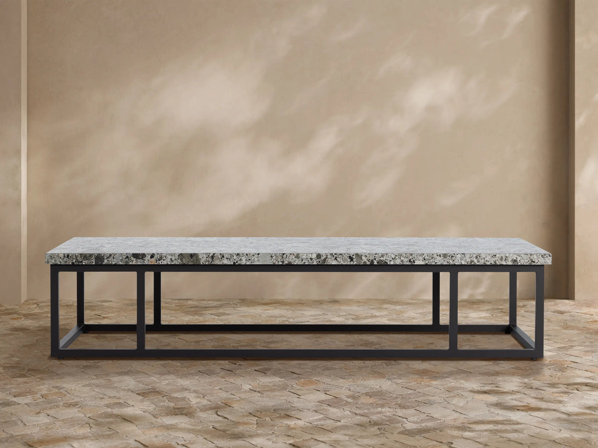 Paloma Outdoor Coffee Table with light gray speckled sintered stone top on a black rectangular metal frame, against a textured tan wall.
