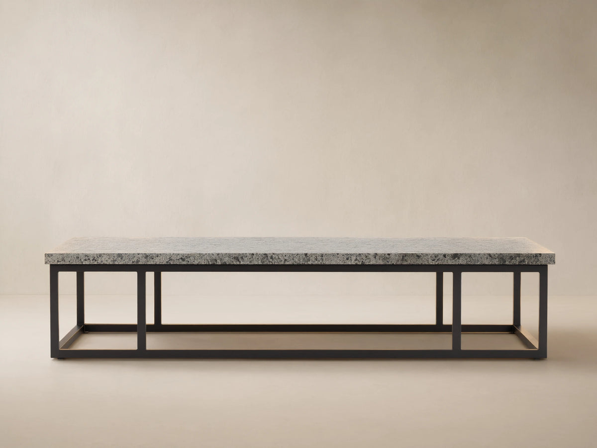 Paloma Outdoor Coffee Table with light gray speckled sintered stone top on a black rectangular metal frame, against a textured tan wall.