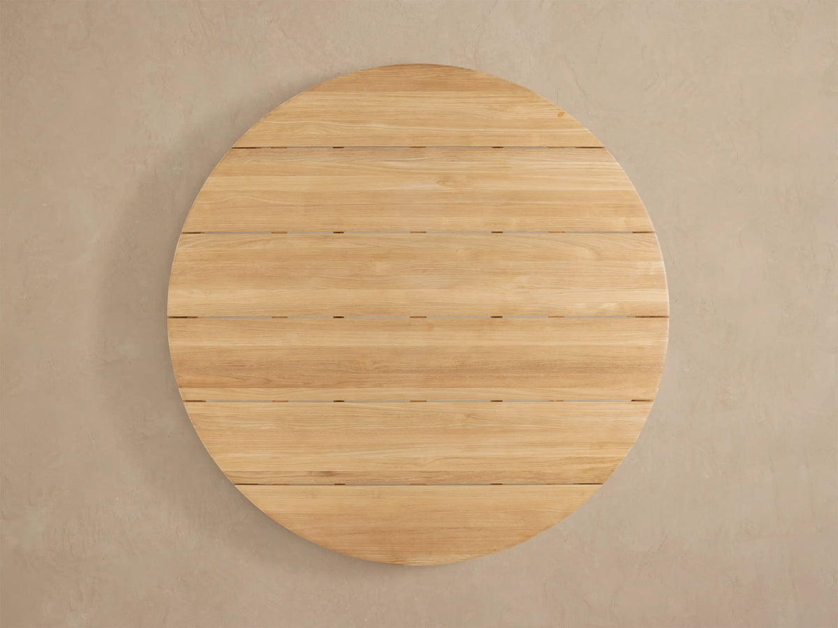 Detail of Puglia Outdoor Teak Round Dining Table. Golden, unfinished teak wood with visible grain, curved edge, and leg.