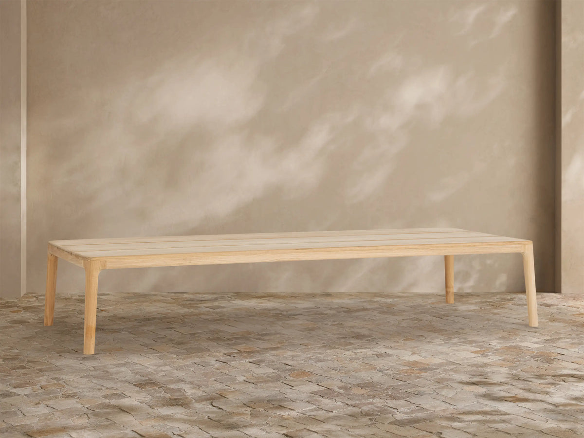 Puglia Outdoor Teak Coffee Table, light golden untreated teak wood, rectangular slatted top, sturdy legs, on stone patio.