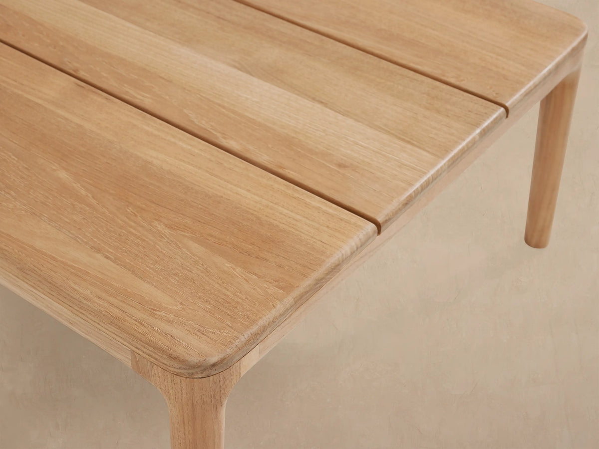 Puglia Outdoor Teak Coffee Table: A long, rectangular, slatted table of light natural teak wood on a stone patio.