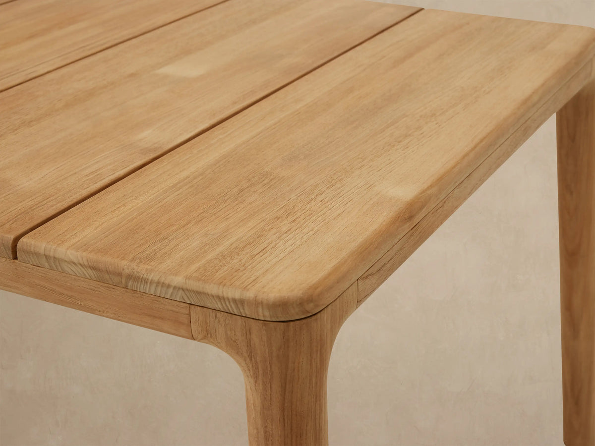 Puglia Outdoor Teak Dining Table: rectangular light natural teak wood table with slatted top and sturdy legs on a stone patio.