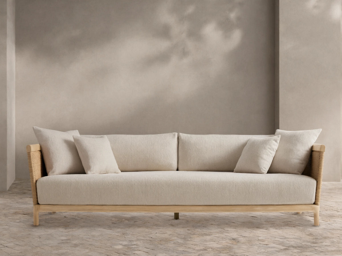 Puglia Outdoor Teak Sofa, light-toned wood frame, woven sides, cream cushions & pillows, on a patterned patio floor before a beige wall.