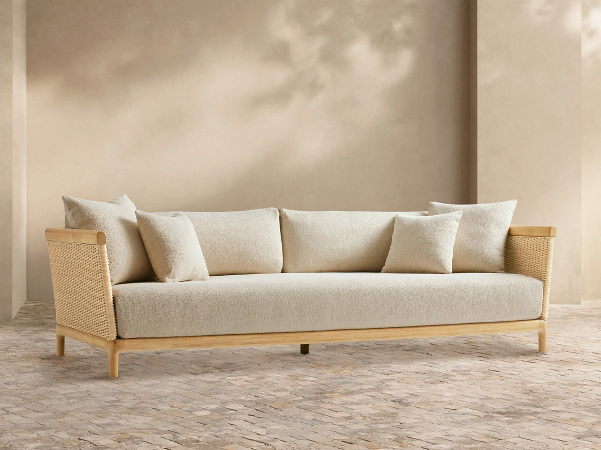 Puglia Outdoor Teak Sofa, light-toned wood frame, woven sides, cream cushions & pillows, on a patterned patio floor before a beige wall.