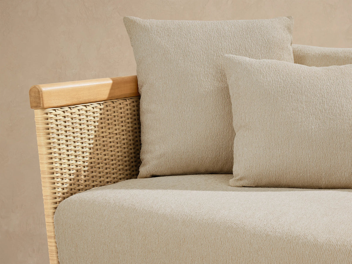Puglia Outdoor Teak Sofa with light beige cushions & throw pillows, natural teak frame, and woven wicker side panels on a patio.