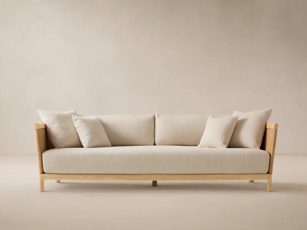 Puglia Outdoor Teak Sofa, light-toned wood frame, woven sides, cream cushions & pillows, on a patterned patio floor before a beige wall.