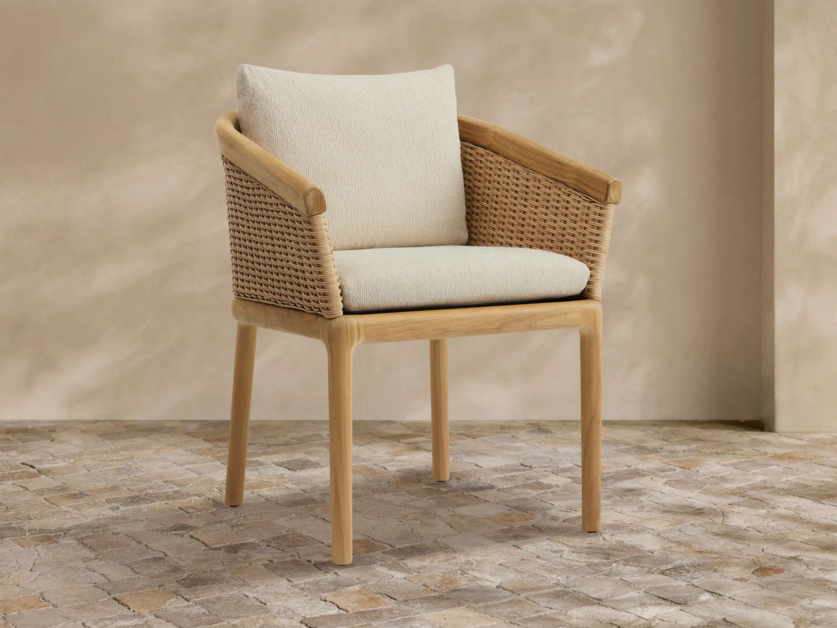 Puglia Outdoor Teak Dining Chair with woven wicker back and armrests, light teak frame, and cream cushions on a stone patio.