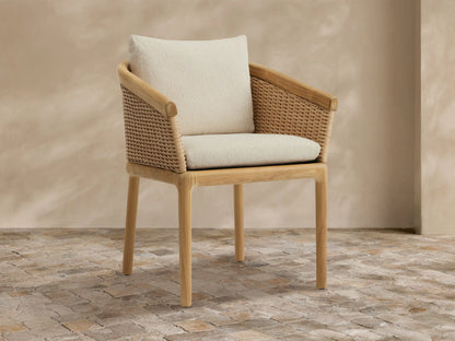 Puglia Outdoor Teak Dining Chair with woven wicker back and armrests, light teak frame, and cream cushions on a stone patio.