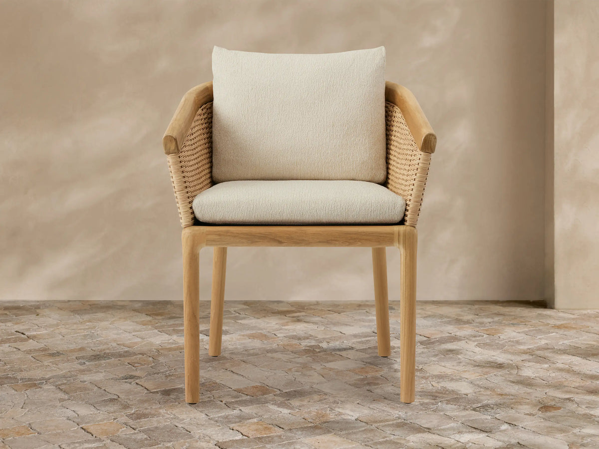 Puglia Outdoor Teak Dining Chair with woven wicker back and armrests, light teak frame, and cream cushions on a stone patio.