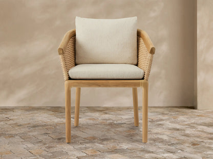 Puglia Outdoor Teak Dining Chair with woven wicker back and armrests, light teak frame, and cream cushions on a stone patio.