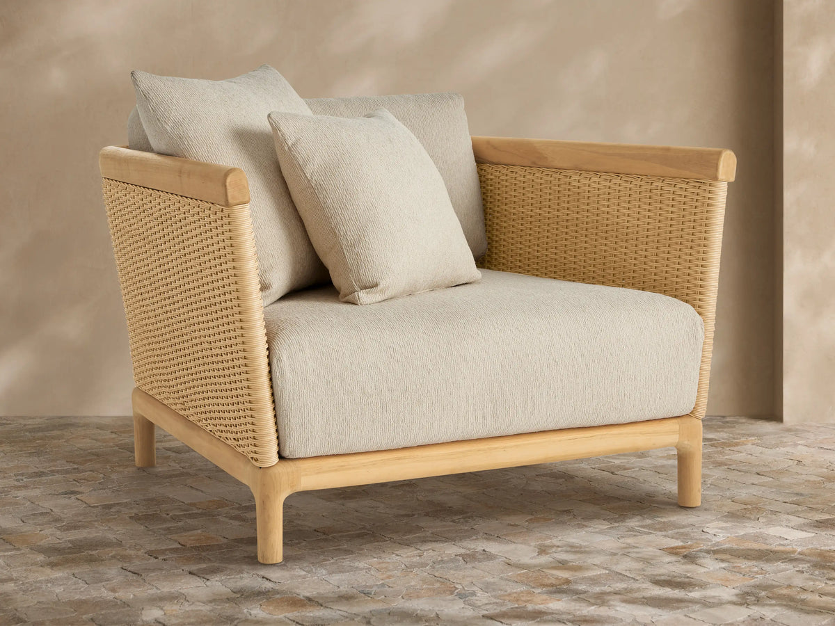 Puglia Outdoor Teak Lounge Chair with natural teak frame, woven tan wicker sides, and light beige cushions, angled right on a stone patio.