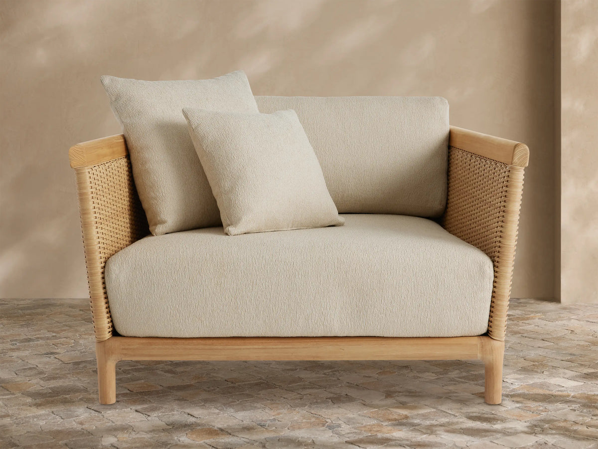 Angled Puglia Outdoor Teak Lounge Chair with natural teak frame, woven wicker sides, and plush cream cushions on a stone patio.