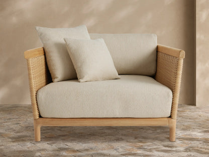 Angled Puglia Outdoor Teak Lounge Chair with natural teak frame, woven wicker sides, and plush cream cushions on a stone patio.