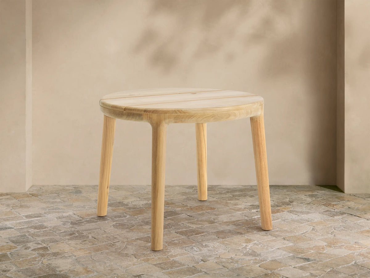 Round Puglia Outdoor Teak End Table with light natural wood, slatted top & four cylindrical legs, on rustic tiled patio.