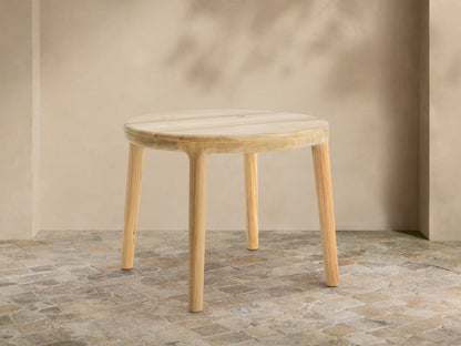 Round Puglia Outdoor Teak End Table with light natural wood, slatted top & four cylindrical legs, on rustic tiled patio.
