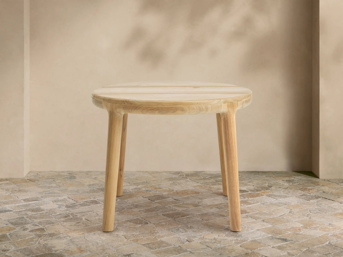 Round Puglia Outdoor Teak End Table with light natural wood, slatted top & four cylindrical legs, on rustic tiled patio.