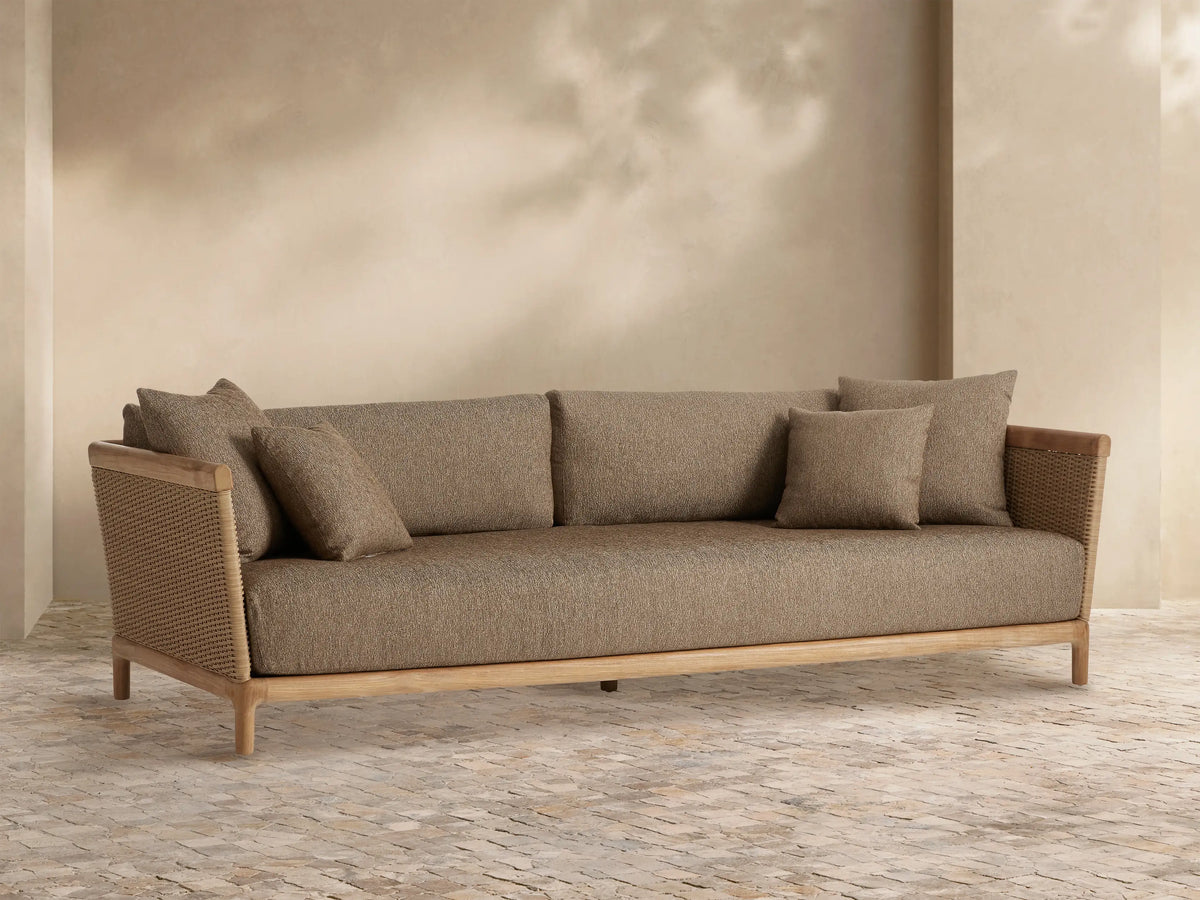 Front-facing Puglia Outdoor Teak Sofa with light brown teak frame, woven sides, and dark tan cushions on an outdoor patio.