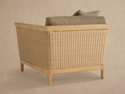 Close-up of Puglia Outdoor Teak Lounge Chair. Light teak wood armrest, woven wicker side, and textured brown cushions with a toss pillow.
