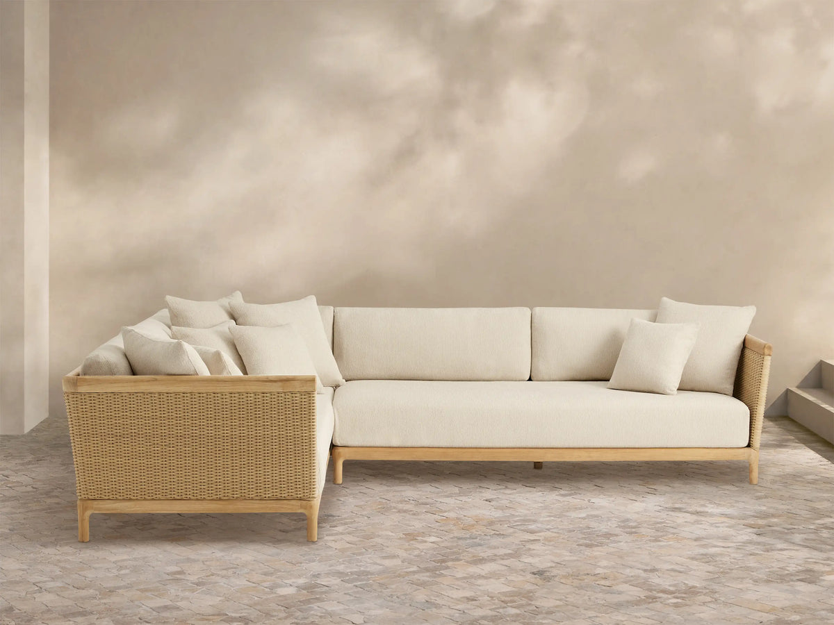 Puglia Outdoor Teak L-shaped Sectional with natural teak frame, light cream cushions, and woven wicker accents on an outdoor patio.