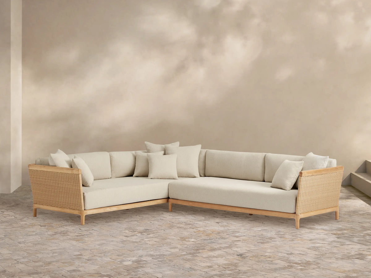 Puglia Outdoor Teak L-shaped Sectional with natural teak frame, light cream cushions, and woven wicker accents on an outdoor patio.