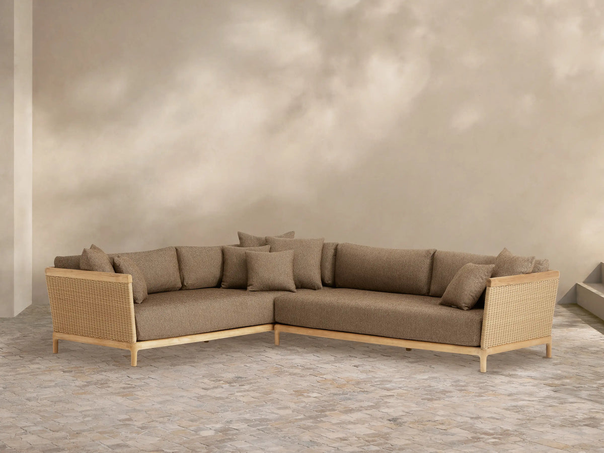L-shaped Puglia Outdoor Teak Two-Piece Sectional with natural teak frame, woven wicker sides, and light brown cushions on a stone patio.