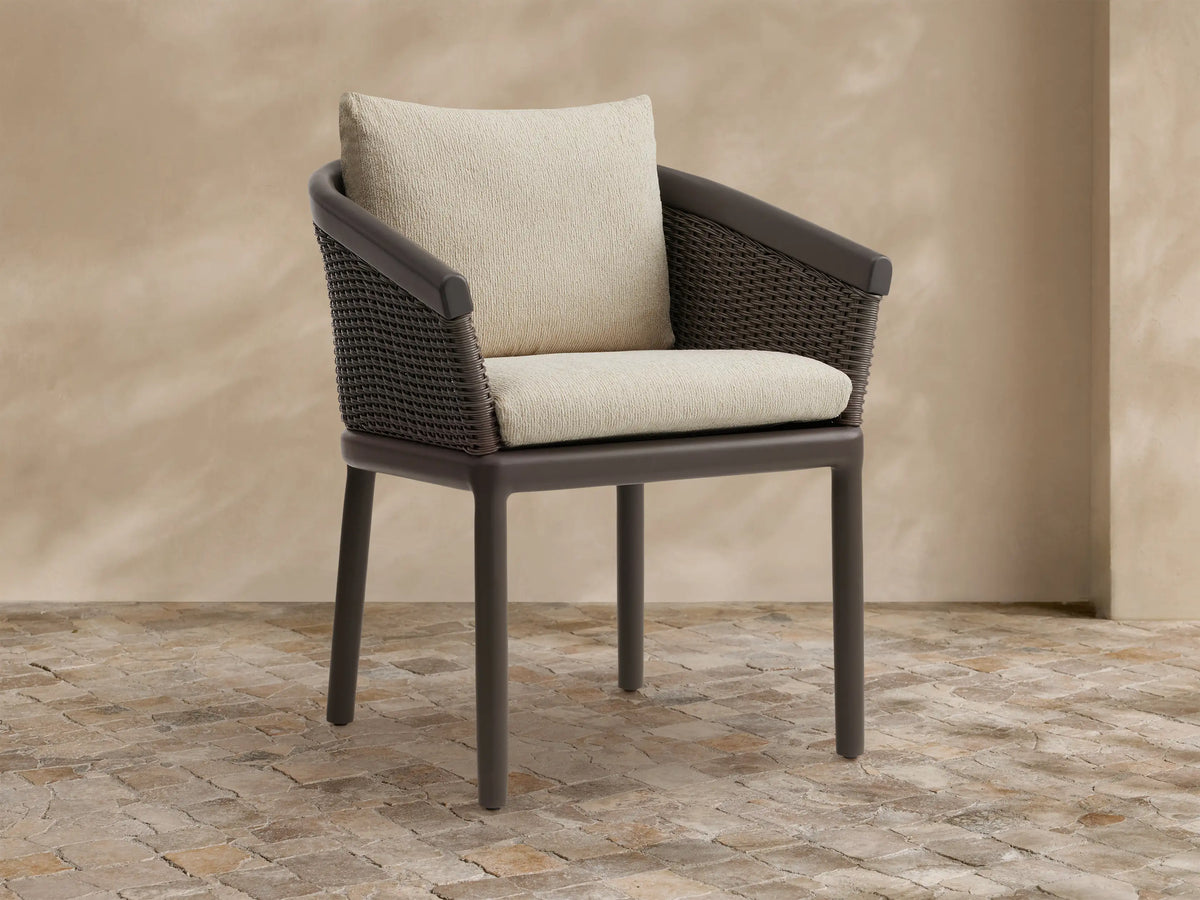 Puglia Outdoor Aluminum Dining Chair with dark brown frame & woven back, light cream seat & back cushions, on stone.
