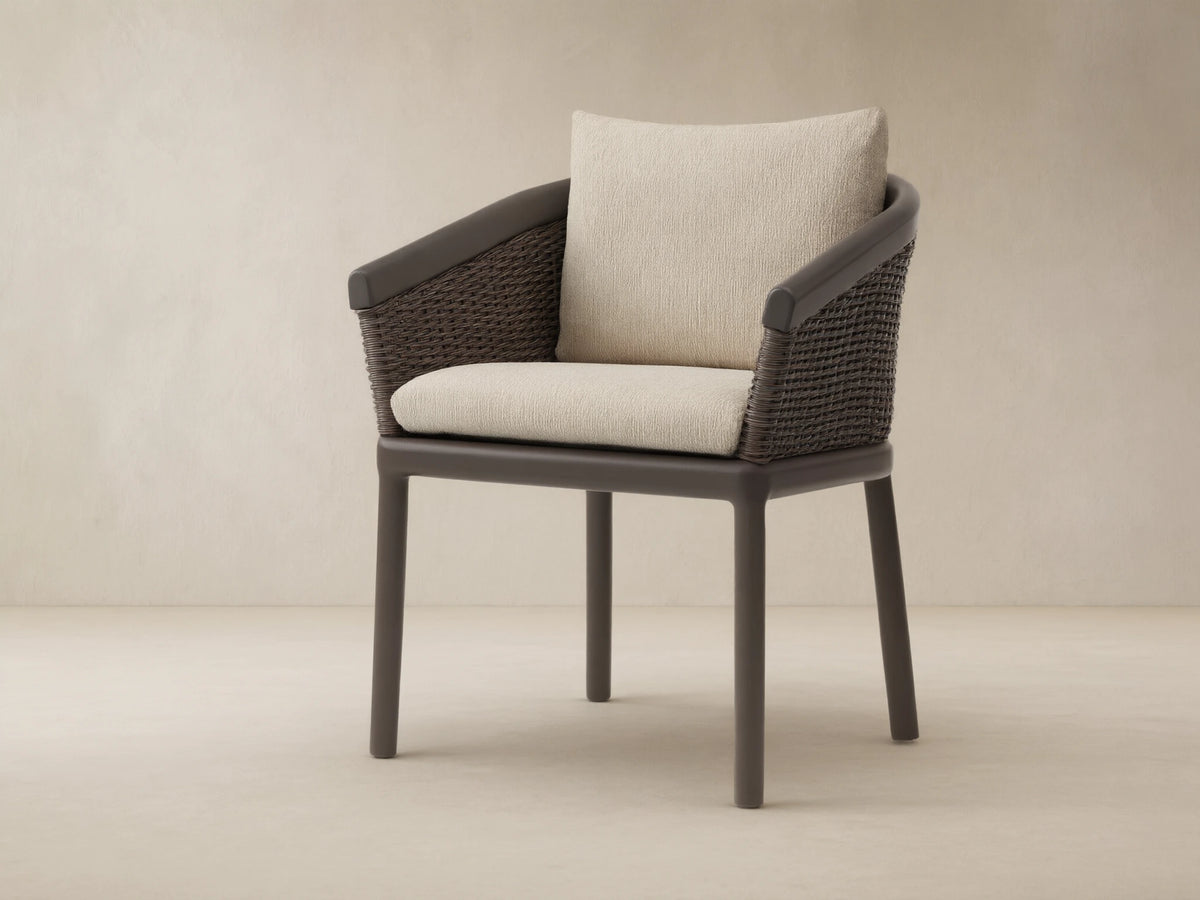 Puglia Outdoor Aluminum Dining Chair with dark brown frame & woven back, light cream seat & back cushions, on stone.