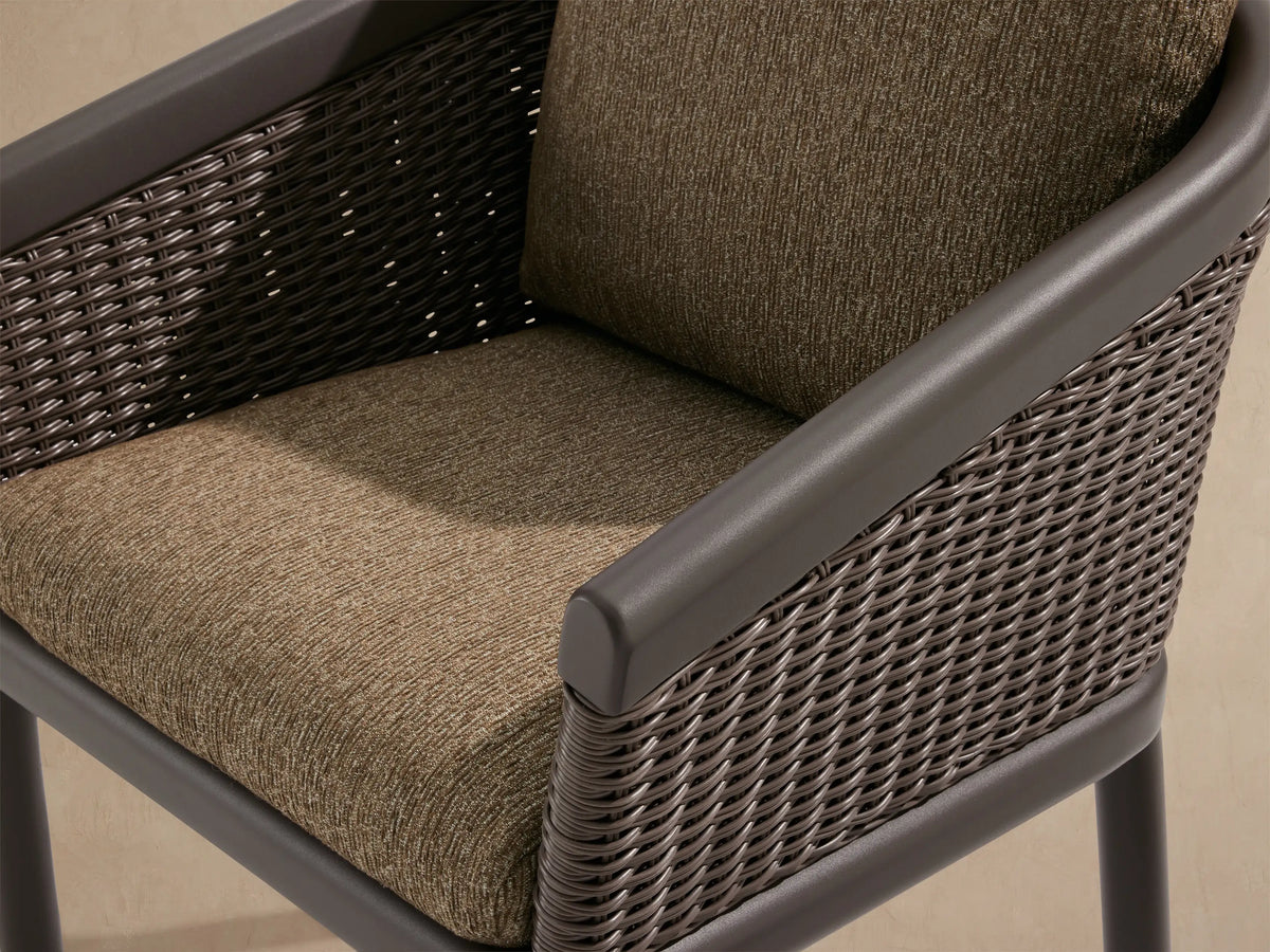Puglia Outdoor Aluminum Dining Chair, front. Dark gray frame, woven resin wicker back & arms, with light brown textured cushions on stone patio.