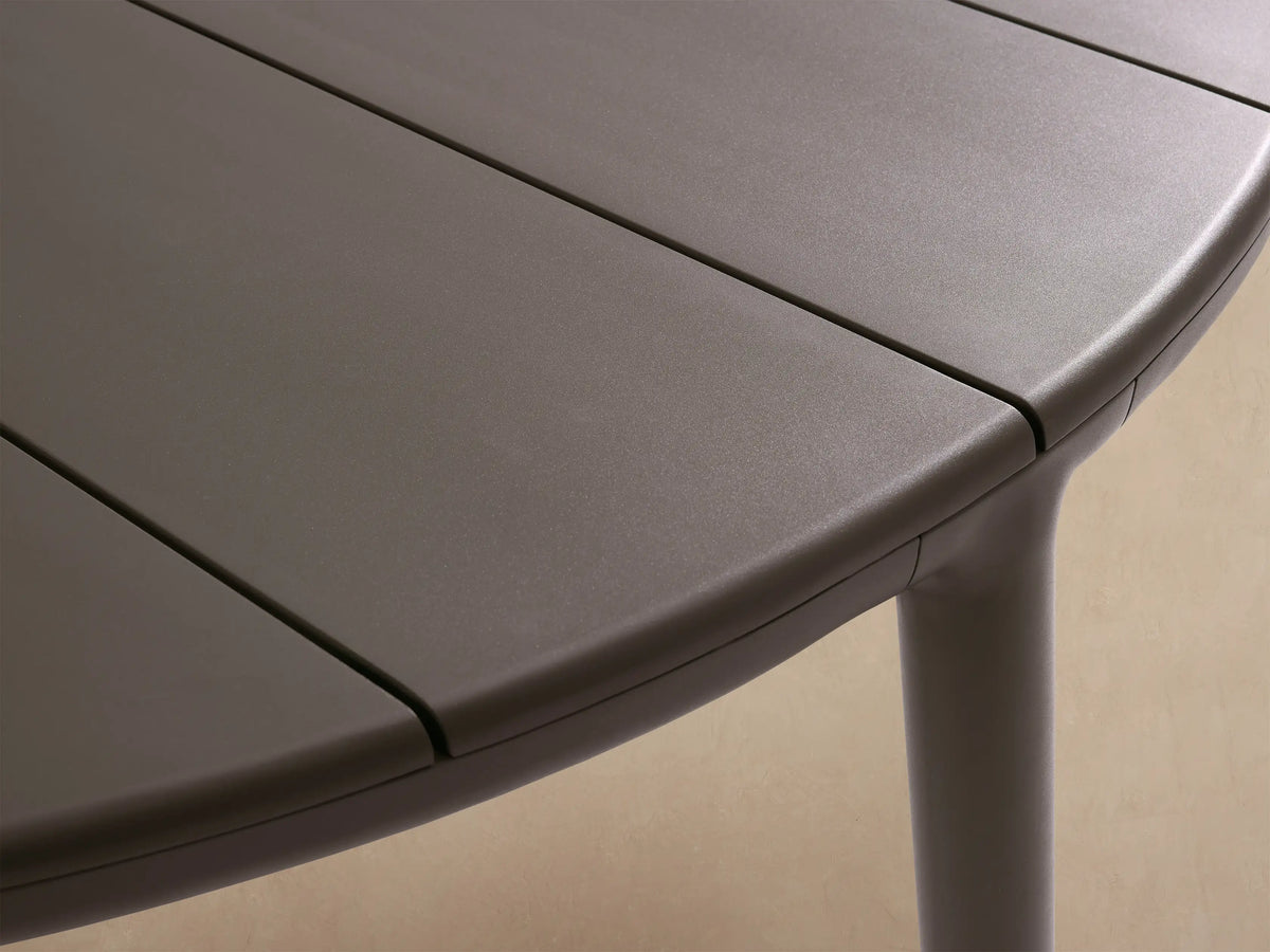 Puglia Outdoor Aluminum Round Dining Table in dark gray, with a slatted top and four sturdy legs, on a patterned stone patio.