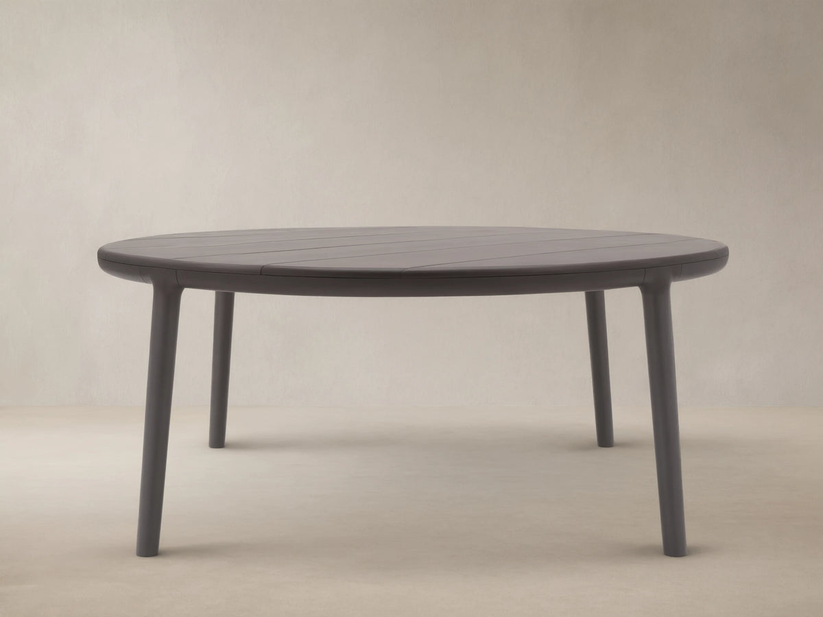 Puglia Outdoor Aluminum Round Dining Table in dark gray, with a slatted top and four sturdy legs, on a patterned stone patio.