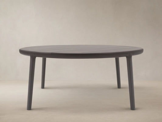 Puglia Outdoor Aluminum Round Dining Table in dark gray, with a slatted top and four sturdy legs, on a patterned stone patio.