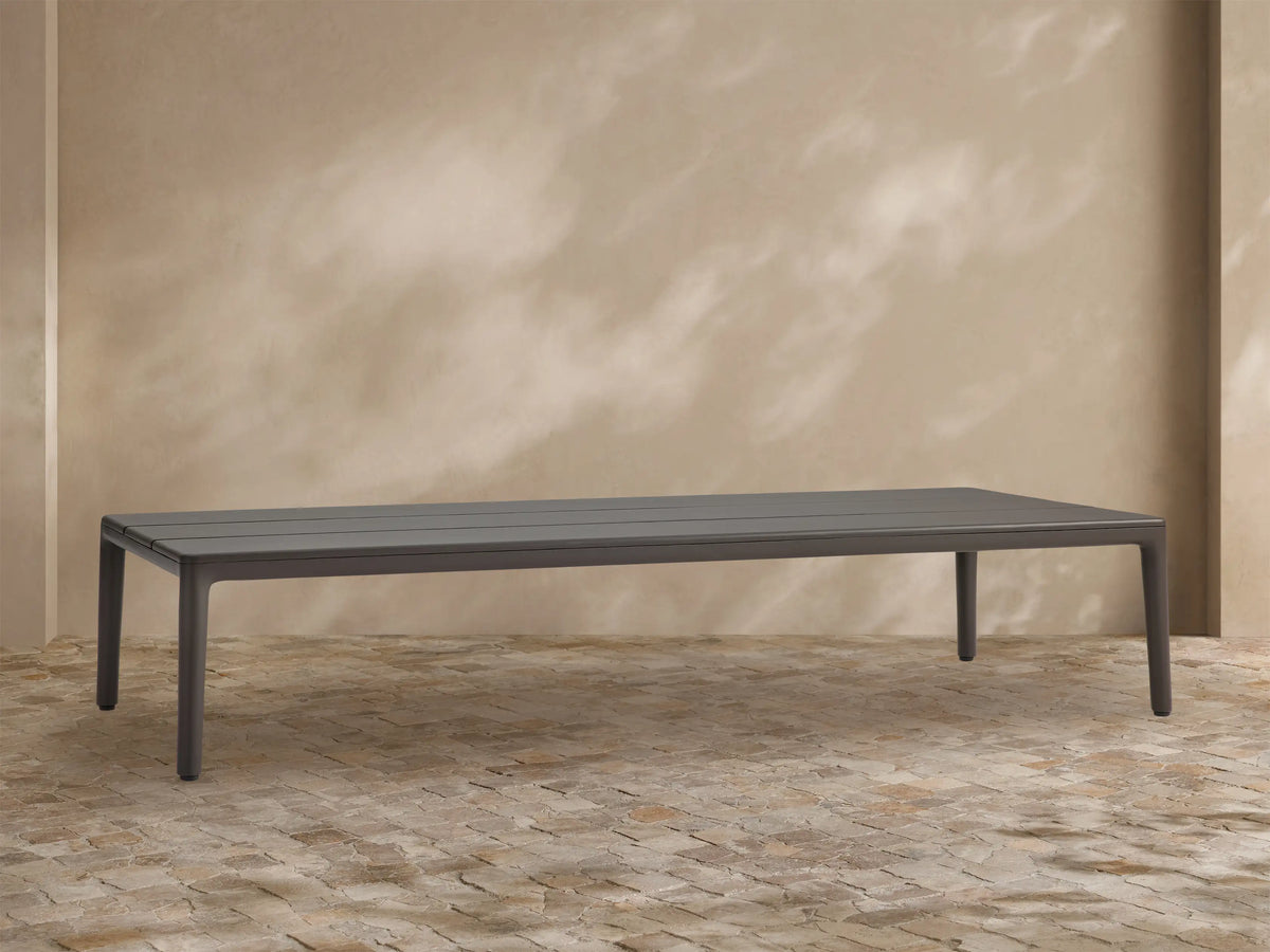 Rectangular dark grey Puglia Outdoor Aluminum Coffee Table with a slatted top, shown outdoors on a light brown textured patio.
