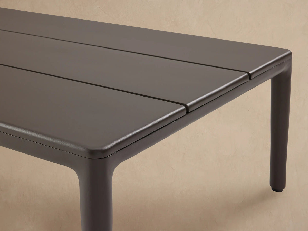 Puglia Outdoor Aluminum Coffee Table, dark gray rectangular with a slatted top and four legs, sitting on a textured stone patio.