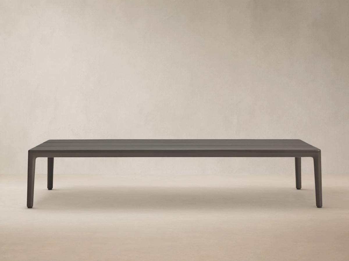 Rectangular dark grey Puglia Outdoor Aluminum Coffee Table with a slatted top, shown outdoors on a light brown textured patio.