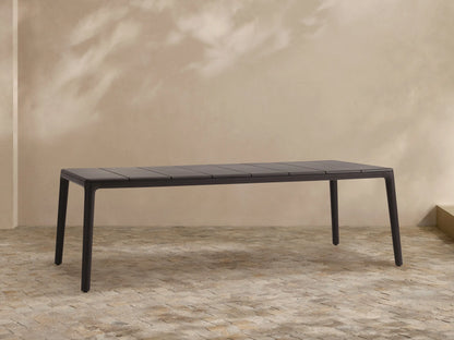 Dark gray Puglia Outdoor Aluminum Dining Table with a slatted top and straight legs, set on a textured patio against a light wall.