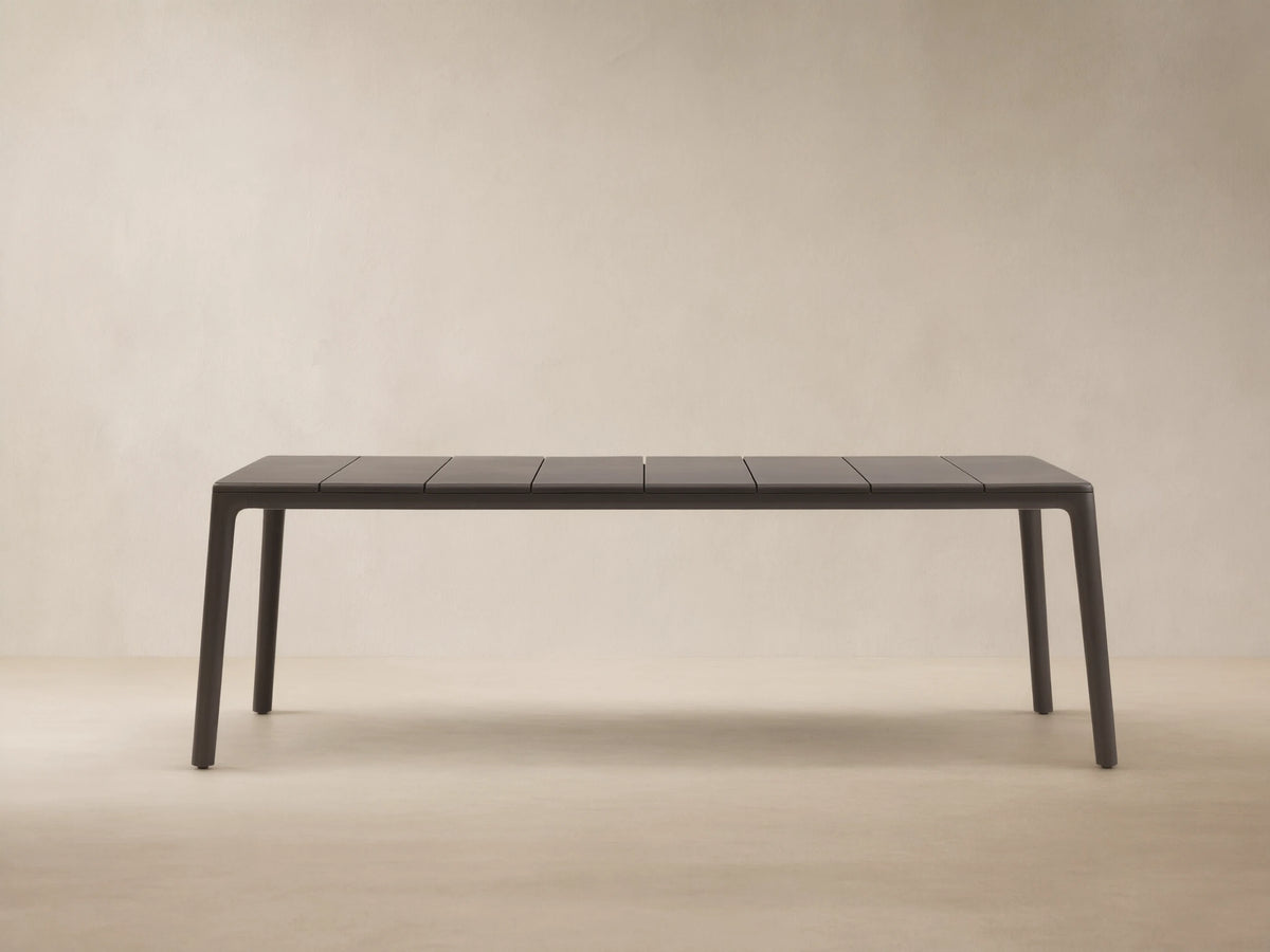 Dark gray Puglia Outdoor Aluminum Dining Table with a slatted top and straight legs, set on a textured patio against a light wall.