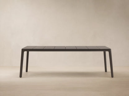 Dark gray Puglia Outdoor Aluminum Dining Table with a slatted top and straight legs, set on a textured patio against a light wall.