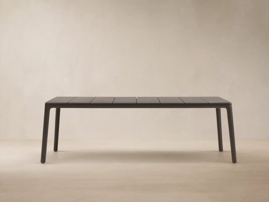 Dark gray Puglia Outdoor Aluminum Dining Table with a slatted top and straight legs, set on a textured patio against a light wall.