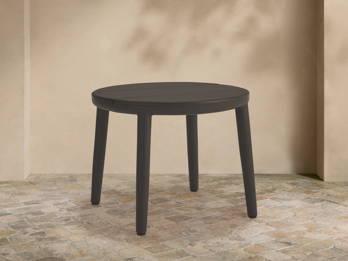 Round dark gray Puglia Outdoor Aluminum End Table, slatted top, four legs, on stone patio with beige wall.