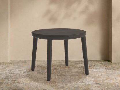 Round dark gray Puglia Outdoor Aluminum End Table, slatted top, four legs, on stone patio with beige wall.