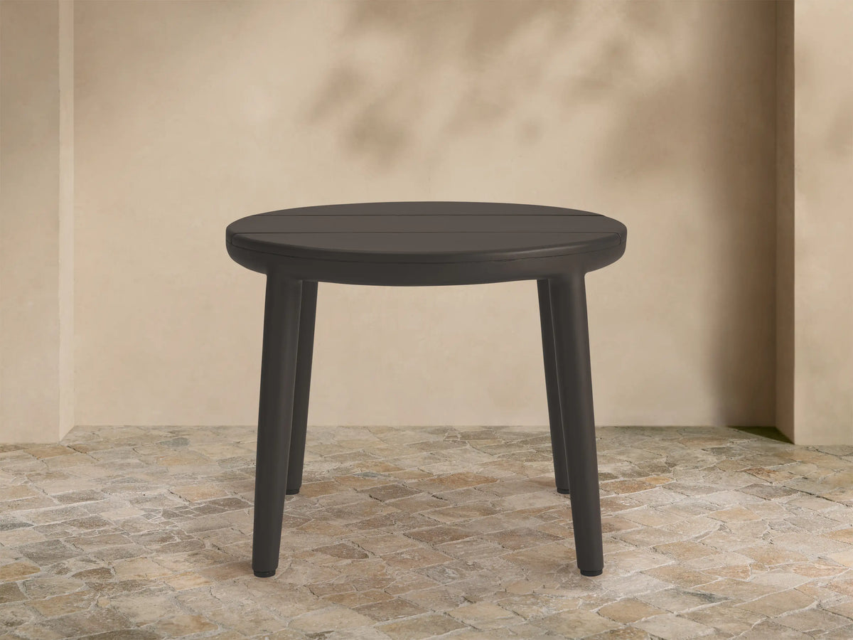 Round dark gray Puglia Outdoor Aluminum End Table, slatted top, four legs, on stone patio with beige wall.