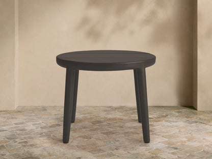 Round dark gray Puglia Outdoor Aluminum End Table, slatted top, four legs, on stone patio with beige wall.