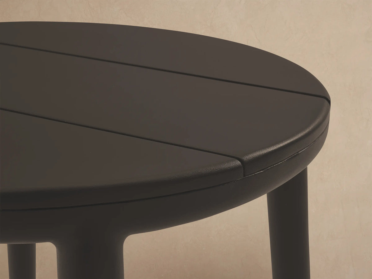 Charcoal gray Puglia Outdoor Aluminum End Table with a slatted round top and four legs on a stone patio by a light wall.
